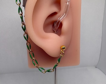 May include: A gold-colored earring with a green chain design is attached to a prosthetic ear. The earring has a small gold ball stud and a chain that connects to a clear plastic hearing aid. The ear is flesh-toned and mounted on a metal stand.