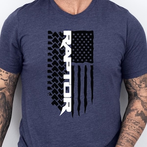 May include: Heather navy V-neck t-shirt with a distressed American flag design and the word "RAPTOR" in white. The flag includes a tire track pattern. The shirt is a dark blue colour.