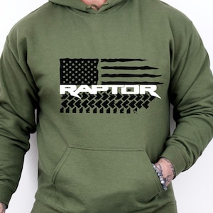Peut inclure: Un sweat à capuche vert avec un motif de drapeau américain noir et blanc et le mot "RAPTOR" en lettres blanches.