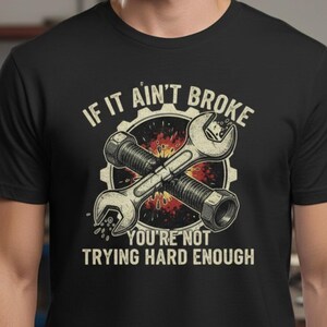 May include: Black t-shirt featuring a graphic of crossed wrenches and a bolt, set against a gear. The text reads "IF IT AIN'T BROKE YOU'RE NOT TRYING HARD ENOUGH" in a vintage style. The shirt is a crew neck.