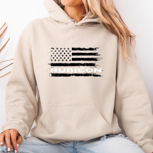 Puede incluir: Una sudadera con capucha beige con un gráfico de bandera estadounidense desgastado en blanco y negro y la palabra "RUBICON" impresa debajo.