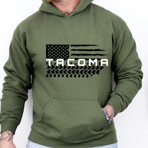 Puede incluir: Una sudadera con capucha verde con un diseño de bandera estadounidense en blanco y negro y la palabra "TACOMA" impresa en blanco.