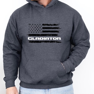 Puede incluir: Una sudadera con capucha gris oscuro con un gráfico de bandera estadounidense desgastado y la palabra "GLADIATOR" impresa en blanco debajo.