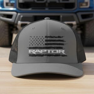 Peut inclure: Casquette de camionneur grise avec un dos en filet noir et une visière incurvée. Le devant présente un motif de drapeau américain vieilli avec le mot "RAPTOR" en blanc. Un camion Ford bleu est visible en arrière-plan.