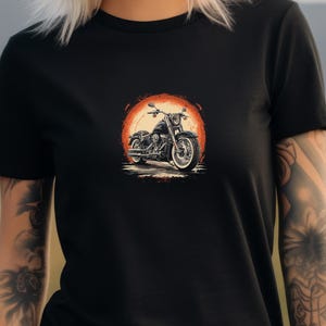 May include: Black t-shirt featuring a motorcycle graphic. The design shows a detailed black motorcycle set against a vibrant orange and red circular background, evoking a sunset or sunrise. The motorcycle has white tires.
