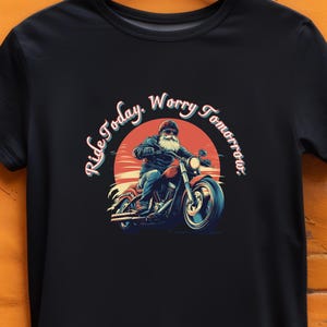May include: Black t-shirt featuring a graphic of Santa Claus riding a red motorcycle against a sunset backdrop. The text "Ride Today, Worry Tomorrow" is arched around the image. The design is a humorous take on the holiday season.