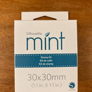 Può includere: Una scatola bianca del kit di timbri Silhouette Mint con la parola "mint" in turchese. La scatola riporta "Kit de sello" e "30x30mm (1,1 pollici x 1,1 pollici)". La scatola è su una superficie di legno.