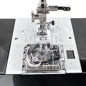 May include: Close-up of a sewing machine's needle and presser foot. The metal plate has measurements in inches, with markings for 1/8, 3/8, and 5/8. The machine is black and silver.