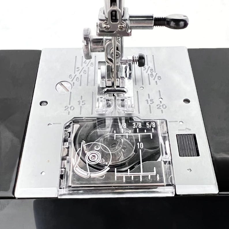 May include: Close-up of a sewing machine's needle and presser foot. The metal plate has measurements in inches, with markings for 1/8, 3/8, and 5/8. The machine is black and silver.