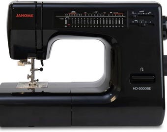 May include: A black Janome HD-5000BE sewing machine. The machine has a variety of stitch options displayed on a panel, with dials and controls on the right side. The Janome logo is in red.