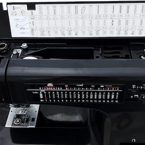 May include: A black sewing machine with a hinged top revealing stitch options. The machine has a control panel with numbered settings and stitch patterns. The brand name is visible in red. The machine is on a white surface.