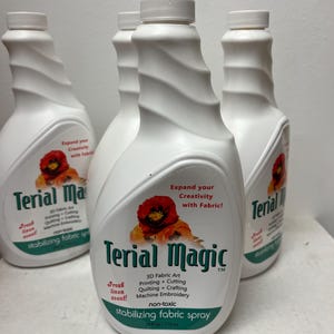 May include: White plastic bottles of Terial Magic stabilizing fabric spray. Each bottle has a white cap and a label with the product name, a red flower graphic, and the text "Expand your Creativity with Fabric!" and "Fresh linen scent!"