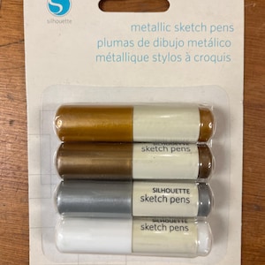 Op de afbeelding: Een verpakking Silhouette metallic schetspotloden. Het pakket bevat vier pennen in goud, brons, zilver en wit. De tekst op de verpakking luidt "metallic sketch pens" en "bevat 4 pennen."