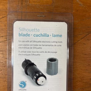 May include: Silhouette blade and ratchet cap in a clear plastic package. The blade is black with white and red markings, and a gray ratchet cap is included. The package text includes "blade cuchilla lame".