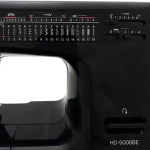May include: Close-up of a black sewing machine with various stitch selection options and control dials. The machine features a range of stitch patterns and settings, with the model number HD-5000BE visible.