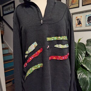 May include: A black long-sleeved shirt with a zippered collar and colorful fabric patches on the front. The patches are made of different fabrics with a variety of patterns.