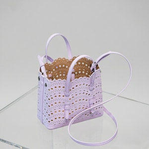 May include: A lavender handbag with a perforated design and a scalloped edge. The bag has two top handles and a detachable shoulder strap. The interior is a light brown color. The bag is sitting on a clear surface.