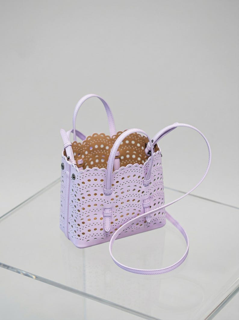 May include: A lavender handbag with a perforated design and a scalloped edge. The bag has two top handles and a detachable shoulder strap. The interior is a light brown color. The bag is sitting on a clear surface.