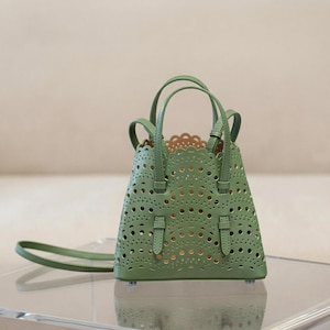 May include: A sage green handbag with a unique cut-out design. The bag features two top handles and a detachable shoulder strap. The interior is a lighter shade, visible through the intricate perforations. The bag has a structured shape and small feet.