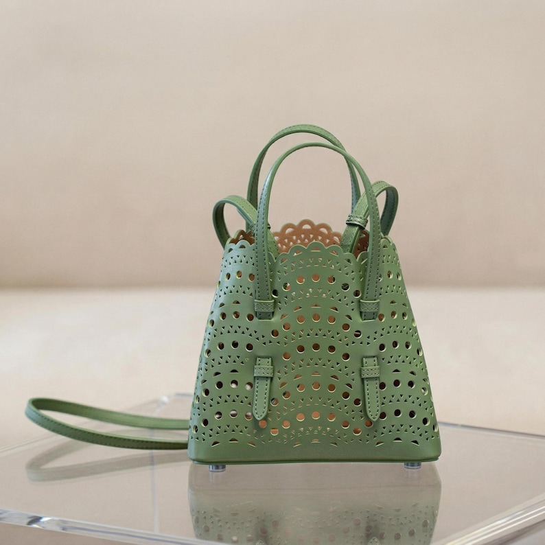 May include: A sage green handbag with a unique cut-out design. The bag features two top handles and a detachable shoulder strap. The interior is a lighter shade, visible through the intricate perforations. The bag has a structured shape and small feet.