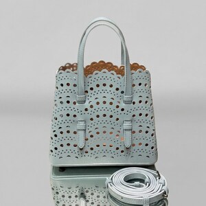 May include: A light blue tote bag with a laser-cut design featuring circular and triangular cutouts. The bag has two top handles and a detachable shoulder strap. The interior is a contrasting tan color.