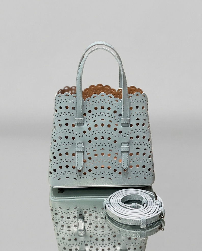 May include: A light blue tote bag with a laser-cut design featuring circular and triangular cutouts. The bag has two top handles and a detachable shoulder strap. The interior is a contrasting tan color.