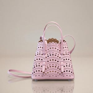 May include: A light pink handbag with a laser-cut design. The bag has two handles and a detachable shoulder strap. The interior is a light brown color. The bag has a rectangular shape with a scalloped top edge and two small buckles.