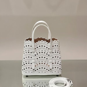 Può includere: Borsa tote bianca tagliata al laser con bordo superiore smerlato e due manici superiori. La borsa è dotata di una tracolla staccabile. L'interno è di colore marrone chiaro. La borsa è appoggiata su una superficie riflettente.