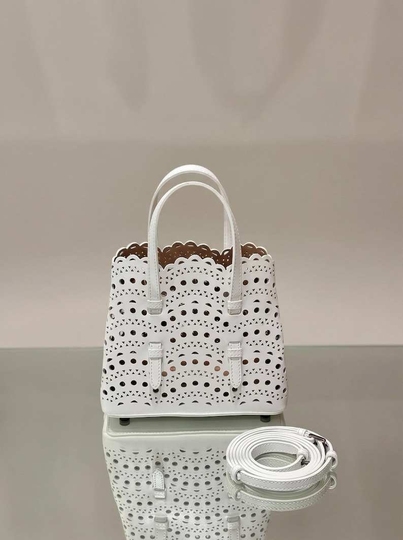 May include: White laser-cut tote bag with a scalloped top edge and two top handles. The bag features a detachable shoulder strap. The interior is a light brown color. The bag is sitting on a reflective surface.