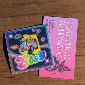 May include: A square sticker with a colorful design featuring two green aliens in a car, a rainbow, and the text "ets9". Next to it is a pink card with the words "ELSEWHERE HANDMADE" and a butterfly graphic.
