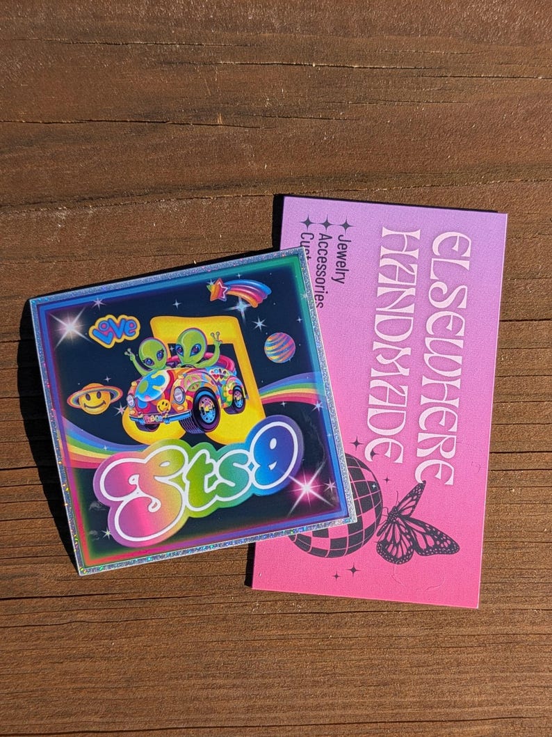 May include: A square sticker with a colorful design featuring two green aliens in a car, a rainbow, and the text "ets9". Next to it is a pink card with the words "ELSEWHERE HANDMADE" and a butterfly graphic.