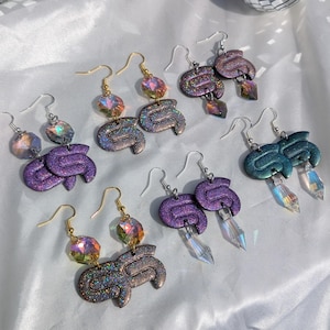 May include: A collection of handmade earrings in various colors, including purple, teal, and iridescent. Each pair features a unique design with glitter accents and crystal or gemstone embellishments. The earrings are displayed on a white surface.