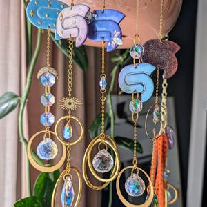 May include: A collection of hanging ornaments featuring sculpted clay shapes in blue, purple, and dark red hues, suspended by gold-toned chains. Each piece incorporates faceted clear crystals, metallic rings, and small sunburst charms.