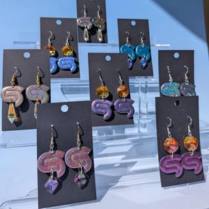 May include: A collection of handmade earrings displayed on black cards. The earrings feature a variety of colors, including purple, blue, and gold, with dangling crystal accents. The earrings have a unique, abstract design.