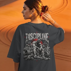 Könnte beinhalten: Ein anthrazitfarbenes T-Shirt mit einem Rückendruck, der das Wort DISCIPLINE in gotischer Schrift zeigt. Das Motiv umfasst eine sitzende Figur, eine Langhantel sowie den Text BUILDS A STRONGER YOU, TODAY NOT TOMORROW und SAME MINDSET STRONGER DAYS.
