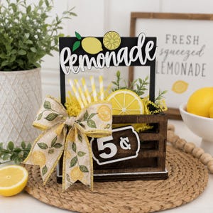 May include: A dark brown wooden crate display featuring a cutout sign that reads lemonade with a lemon graphic. The crate contains yellow paper filler, yellow and white striped straws, and a lemon slice. A burlap bow with a lemon print is attached.