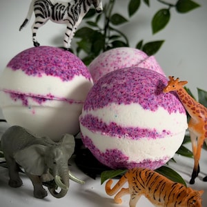 May include: Spherical bath bombs with a white and purple speckled design, and a white stripe. Small animal figurines, such as a zebra, giraffe, elephant, and tiger, are arranged around the bath bombs. Green leaves provide a natural backdrop.
