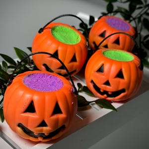 May include: Four orange plastic jack-o'-lantern buckets with black handles and carved faces. The buckets are filled with green and purple material. The buckets are on a white tray with green leaves.