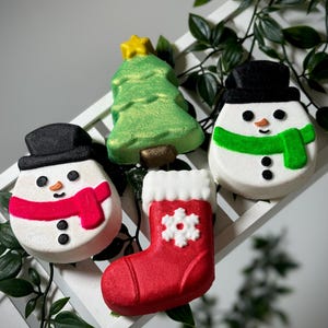 May include: Four festive bath bombs: two snowmen with black hats and scarves, one red, one green. A green Christmas tree with a yellow star and a red stocking with a white snowflake. Festive bath products.