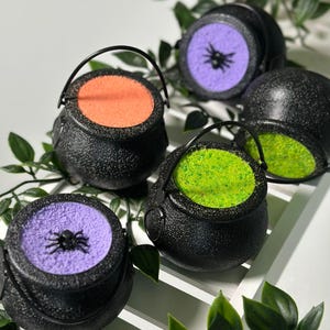 May include: Five black cauldron-shaped containers with handles. Each cauldron has a different coloured, glittery substance inside: orange, green, purple, purple, and purple. The purple cauldrons have a black spider on top.