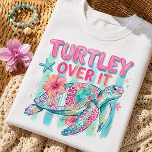 May include: A white t-shirt featuring a colorful graphic of a sea turtle with a patterned shell, pink hibiscus flowers, and a teal starfish. The text "TURTLEY OVER IT" is printed in bold pink letters above the turtle, set against a light blue striped background.