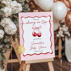 May include: A pink baby shower sign on a wooden easel. The sign features the text "She's the cherry on top" and "Welcome to Samantha's Baby Shower". The sign has a red border and a cherry graphic with a pink bow.