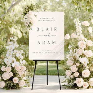 Puede incluir: Un letrero de bienvenida de boda sobre un caballete negro, rodeado de arreglos florales. El letrero dice "Bienvenida a la boda de Blair y Adam" con la fecha y un mensaje de bienvenida. Las flores son rosas blancas y rosadas.