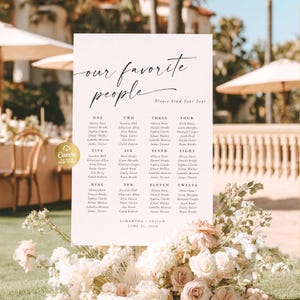 Peut inclure: Un plan de table de mariage avec l'inscription "our favorite people" en écriture cursive. Les noms des invités sont listés sous des numéros de table. Le plan est orné d'une composition florale rose et blanche.