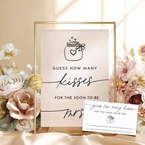 May include: A framed sign and a small card with the text "Guess how many kisses for the soon-to-be Mrs." The sign features a jar with a heart and the word "kisses" in cursive. The background is a soft beige with floral arrangements.
