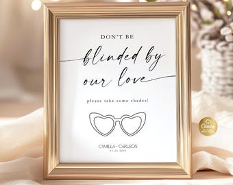 May include: A framed sign with gold trim, featuring the text "DON'T BE blinded by our love" and "please take some shades!" in elegant script. Below is a heart-shaped sunglasses graphic. The sign is for Camilla & Carlson, 05.25.2025.