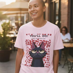 Puede incluir: Camiseta rosa claro con el texto "YOU'LL DO!" y "HAPPY VALENTINE'S DAY". La camiseta presenta una ilustración de un gato negro con acentos de corazón rojo. La persona sonríe y está de pie afuera.