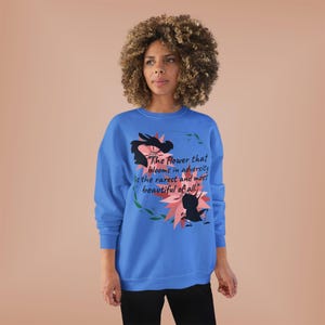 Op de afbeelding: Een blauwe sweatshirt met een bloemenontwerp en de quote: "De bloem die bloeit in tegenspoed is de zeldzaamste en mooiste van allemaal." Het ontwerp bevat roze en zwarte bloemelementen.