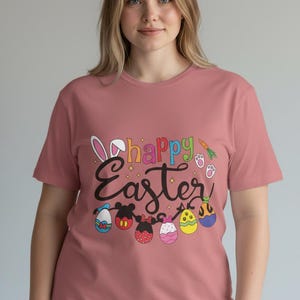Puede incluir: Camiseta rosa polvoriento con las palabras "Happy Easter" en letras coloridas. El diseño incluye orejas de conejo, huevos de Pascua y una zanahoria. La camiseta es de cuello redondo y manga corta.