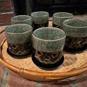 May include: Six ceramic tea cups with a crackle glaze in shades of teal and brown, arranged on a round bamboo tray. Each cup has a unique design and sits on a small black saucer. The tray is on a brick surface.